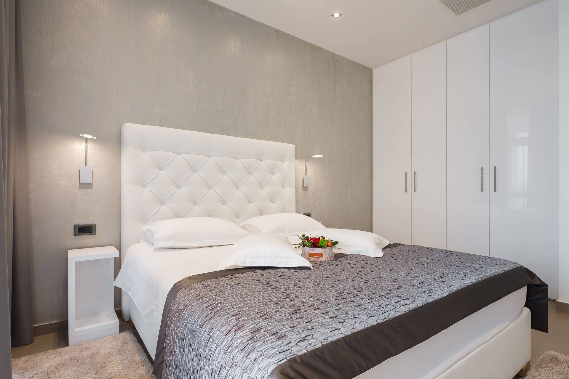 Een moderne slaapkamer met een gestoffeerd hoofdbord, een wit bed, een grijs beddegoed met patroon, en een nachtkastje met een lamp.
