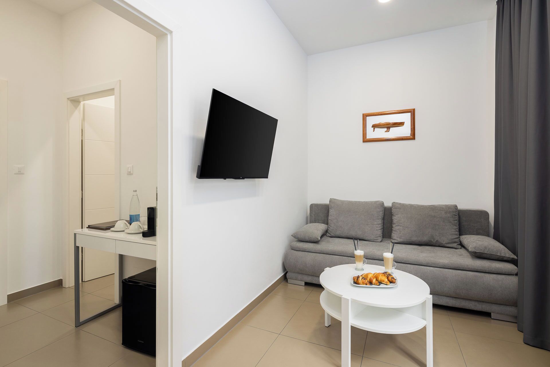 Un salon moderne avec un canapé gris, une petite table blanche sur laquelle se trouve une assiette de croissants, un téléviseur à écran plat fixé au mur et un tableau encadré au-dessus du canapé.
