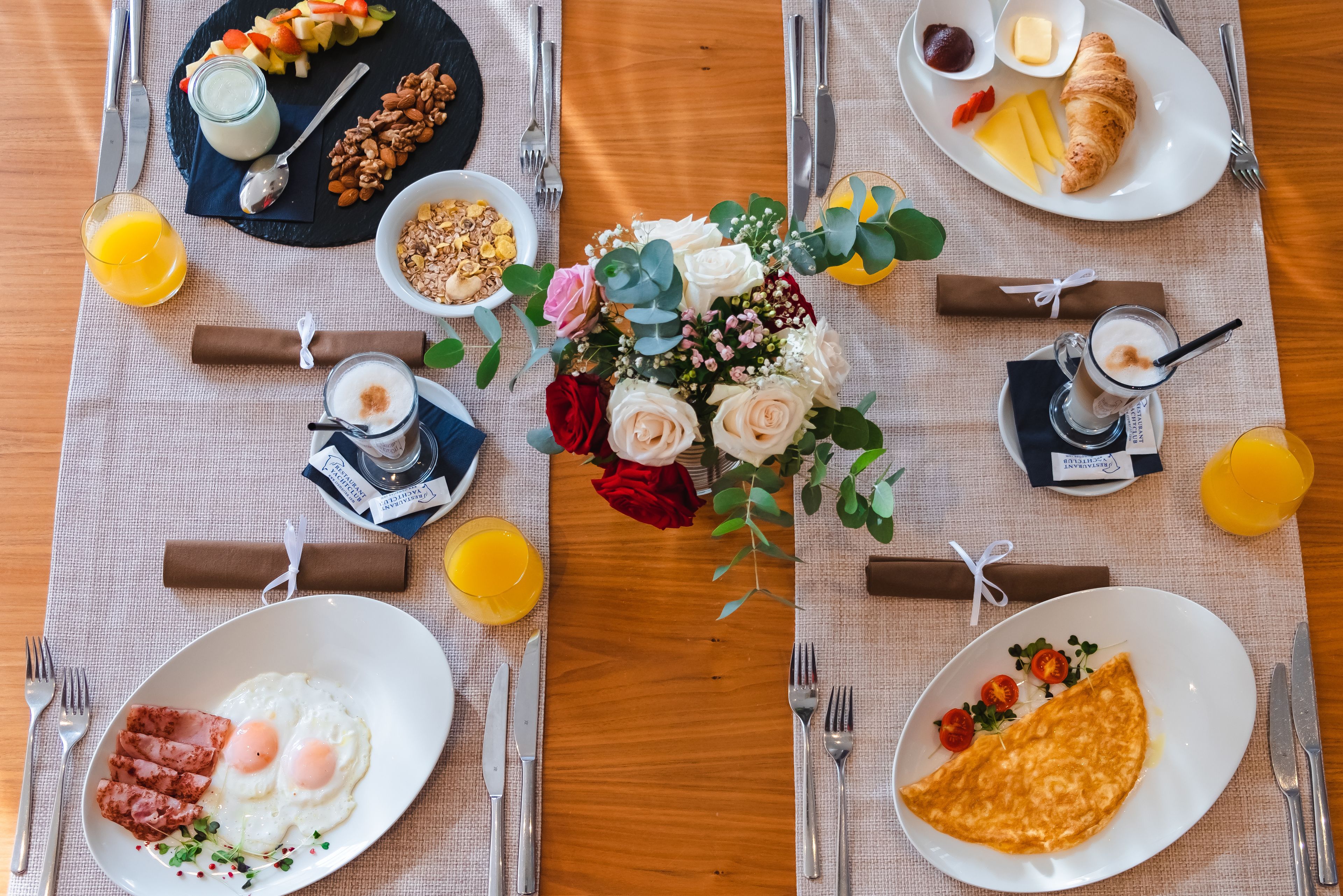 A breakfast setting with plates of eggs, ham, and croissants, a fruit platter, and a vase of flowers.