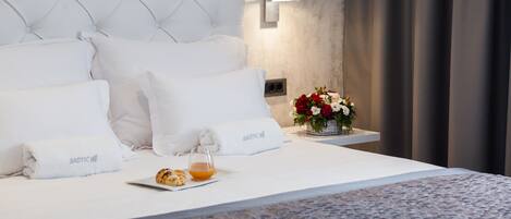 A neatly made bed with white linens and pillows, a small table with a glass of orange juice and croissants, and a vase of red and white flowers on the nightstand.