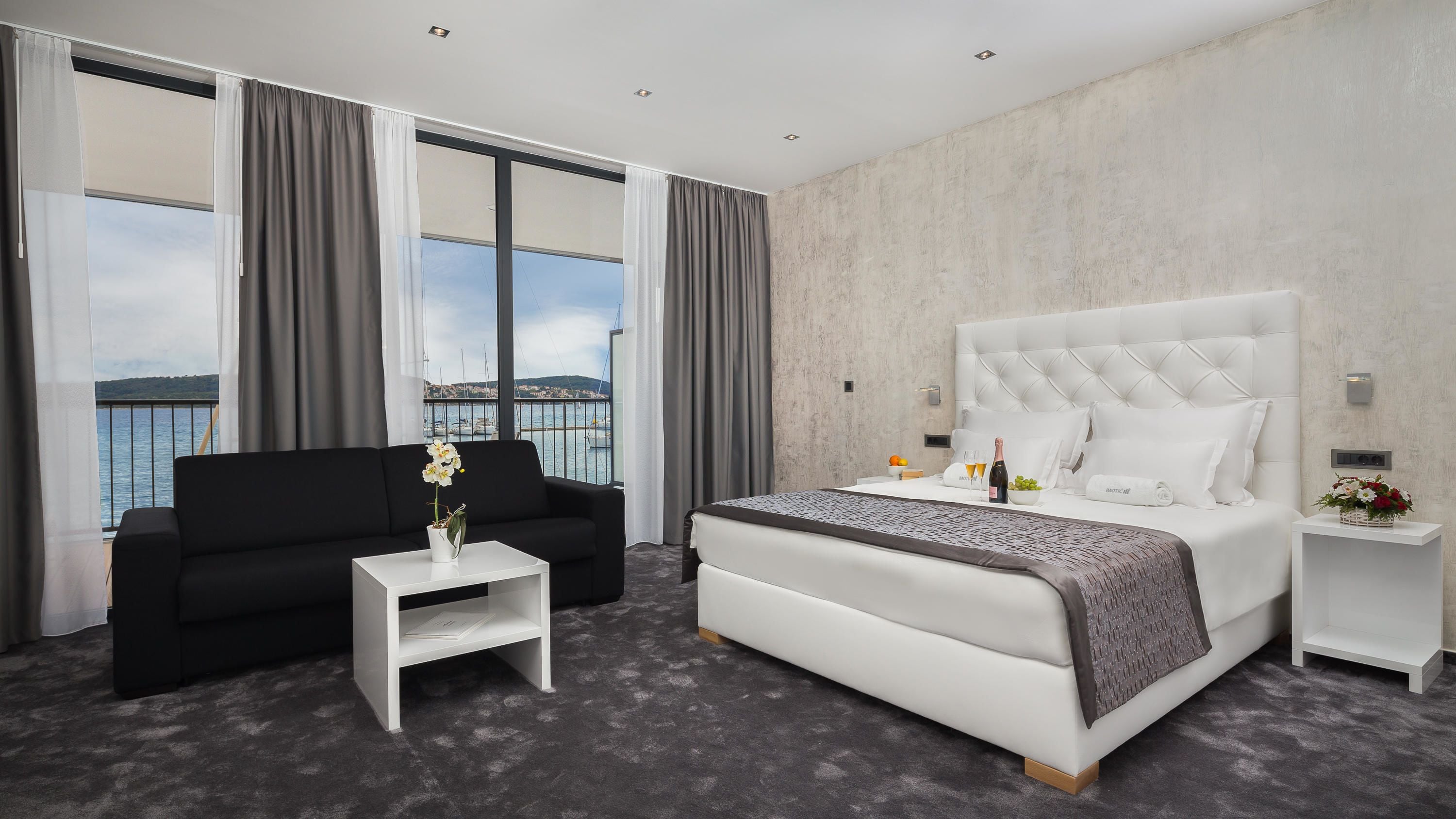 Une chambre d’hôtel moderne avec un grand lit, un canapé noir, une petite table blanche ornée d’un vase de fleurs et une vue sur la mer grâce à de grandes fenêtres.