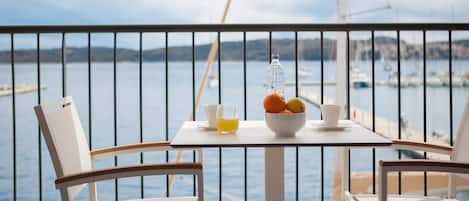 A balcony with a table set for two, featuring a bowl of fruit, a glass of juice, and a bottle of water. The view includes a marina with boats and a distant shoreline.