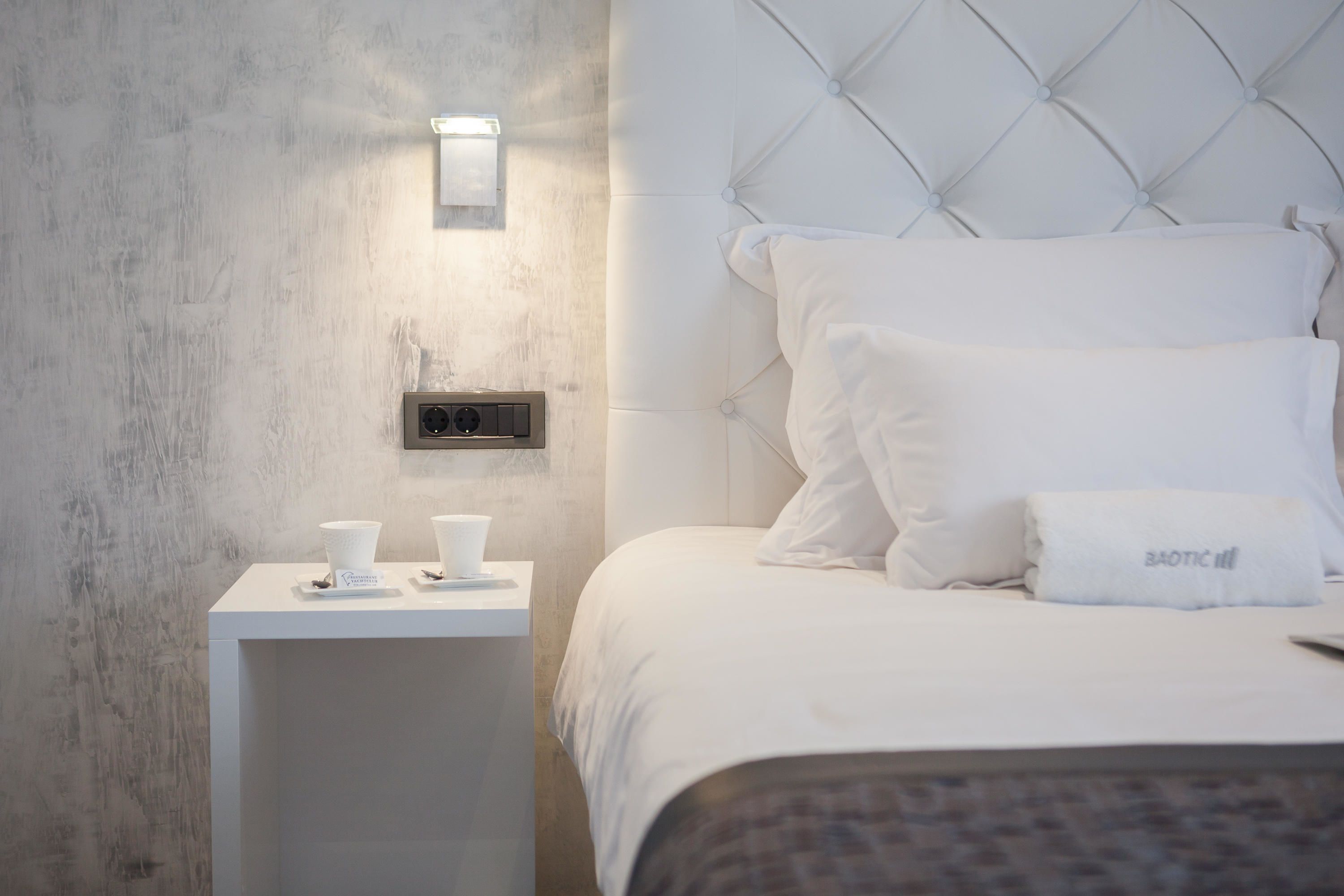 A hotel room with a tufted headboard, a bedside table with a lamp, and a neatly made bed with white linens and a folded towel.