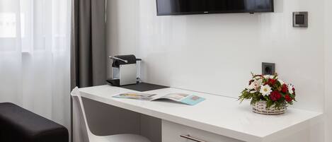 A modern hotel room with a white desk, a black chair, a flat-screen TV mounted on the wall, a small potted plant, and a window with curtains.