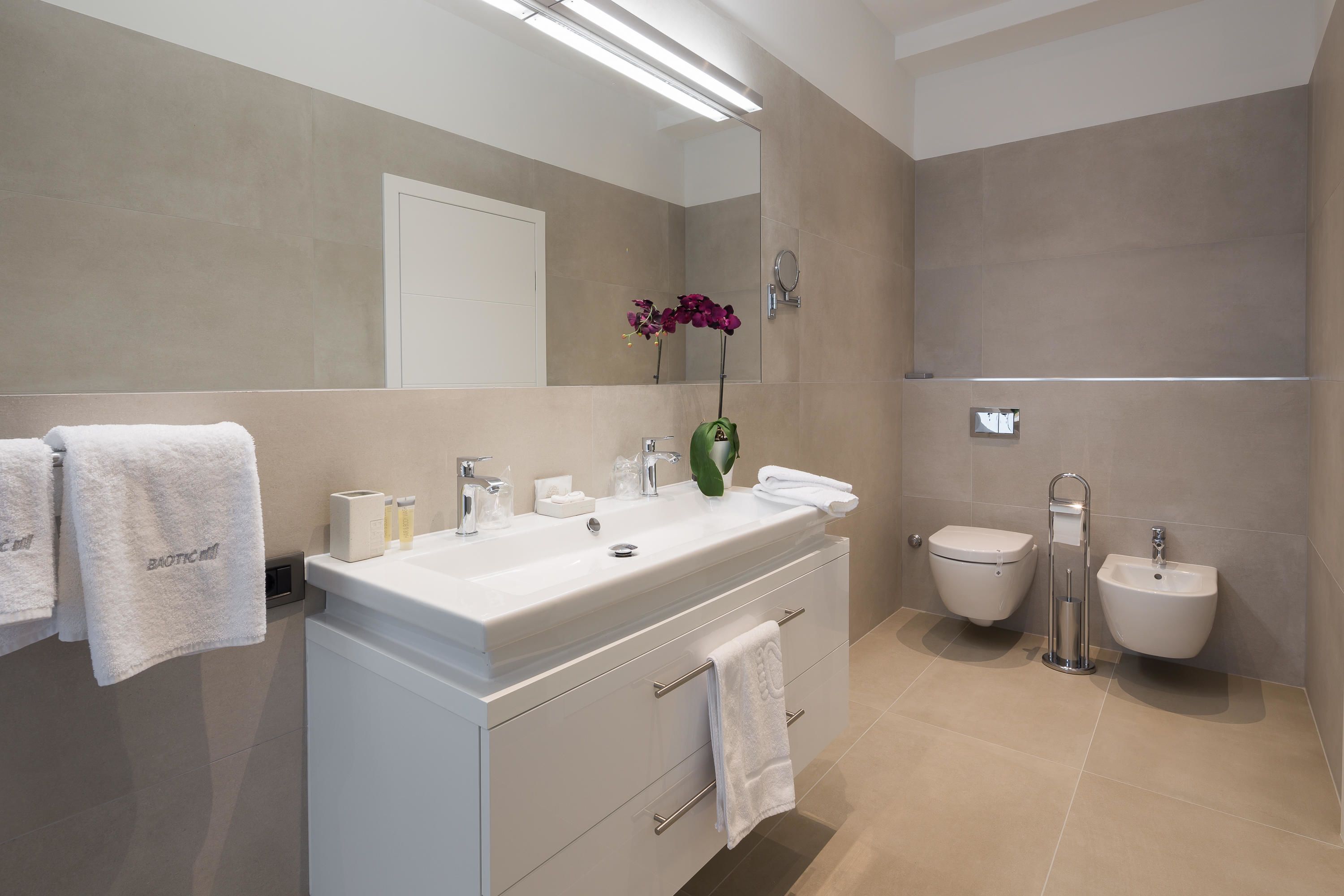 A modern bathroom with a large mirror, a white vanity, a bidet, and a towel rack.