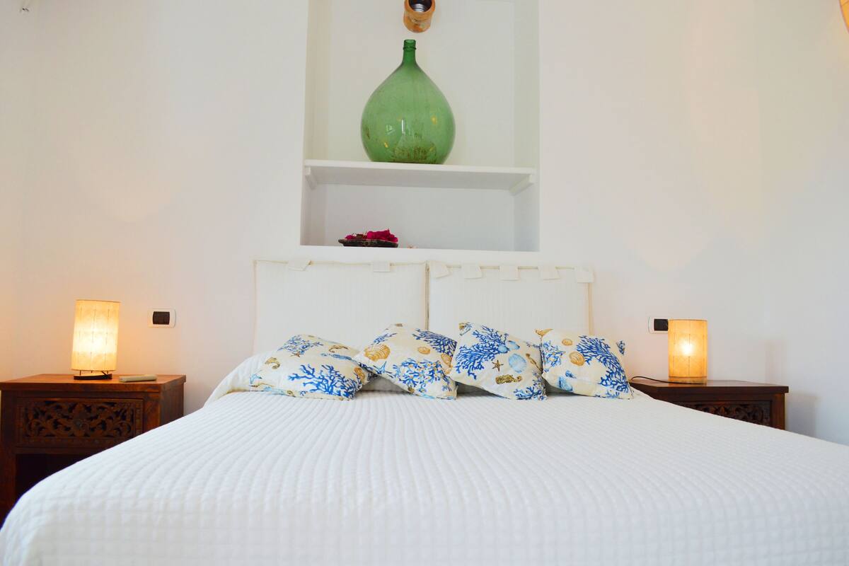 A neatly made bed with white bedding and decorative pillows, two bedside lamps, a green vase on a shelf above the bed, and a small red object on the shelf.