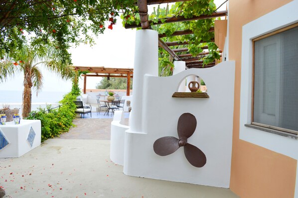 Une terrasse extérieure avec un mur blanc orné d’un grand motif représentant une hélice, une pergola couverte de plantes grimpantes et une vue sur la mer.