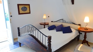 Un dormitorio con una cama de madera, dos mesitas de noche con lámparas, una silla de plástico transparente y un cuadro en la pared.