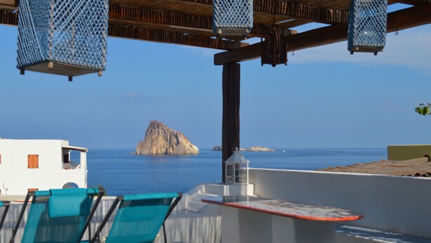 Una terraza con vista al mar, con un edificio blanco, sillas azules y faroles colgantes.