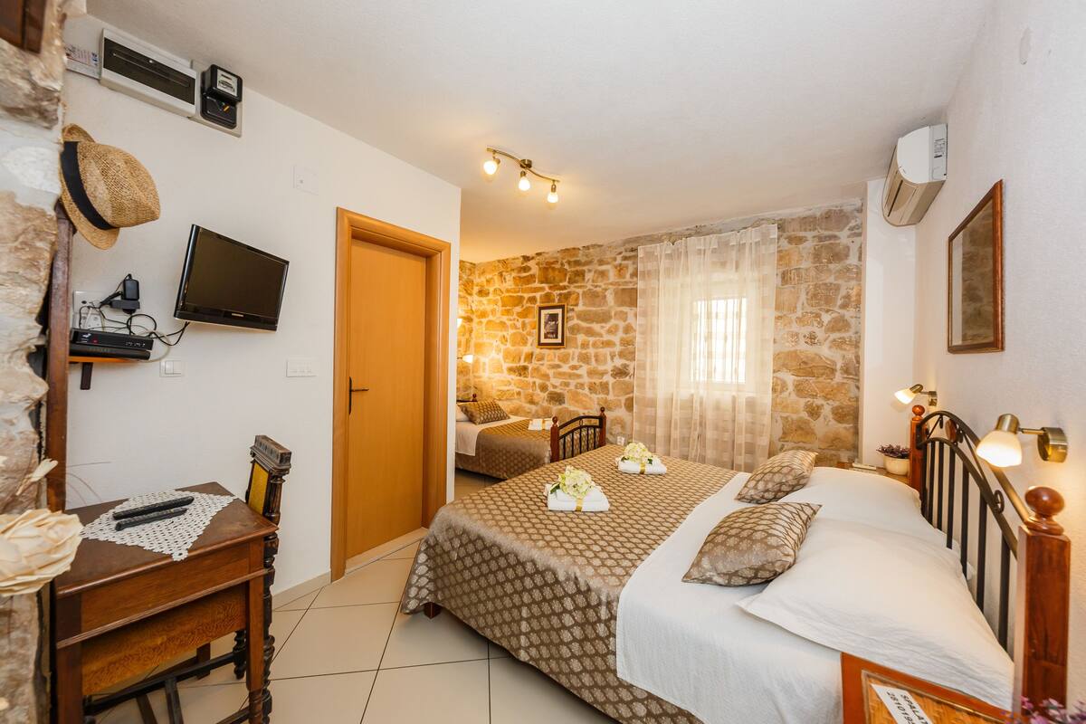 A bedroom with a stone wall, a bed with a patterned bedspread, a bedside table with a remote control, a television mounted on the wall, and a window with sheer curtains.