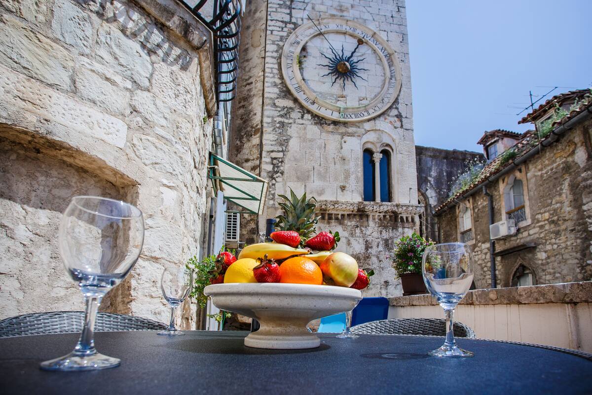 A table with a fruit bowl and two empty wine glasses, set against a stone building with a clock and a window.