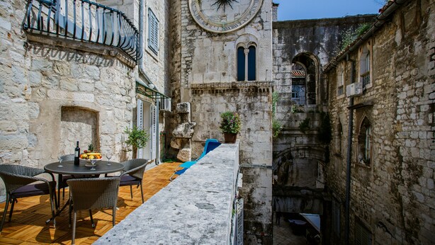 A courtyard with a stone table and chairs, a clock tower, and a narrow alleyway.
