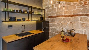 A kitchen with a wooden countertop, a built-in sink, a black refrigerator, and a stone wall. There are shelves above the sink holding dishes and glasses.