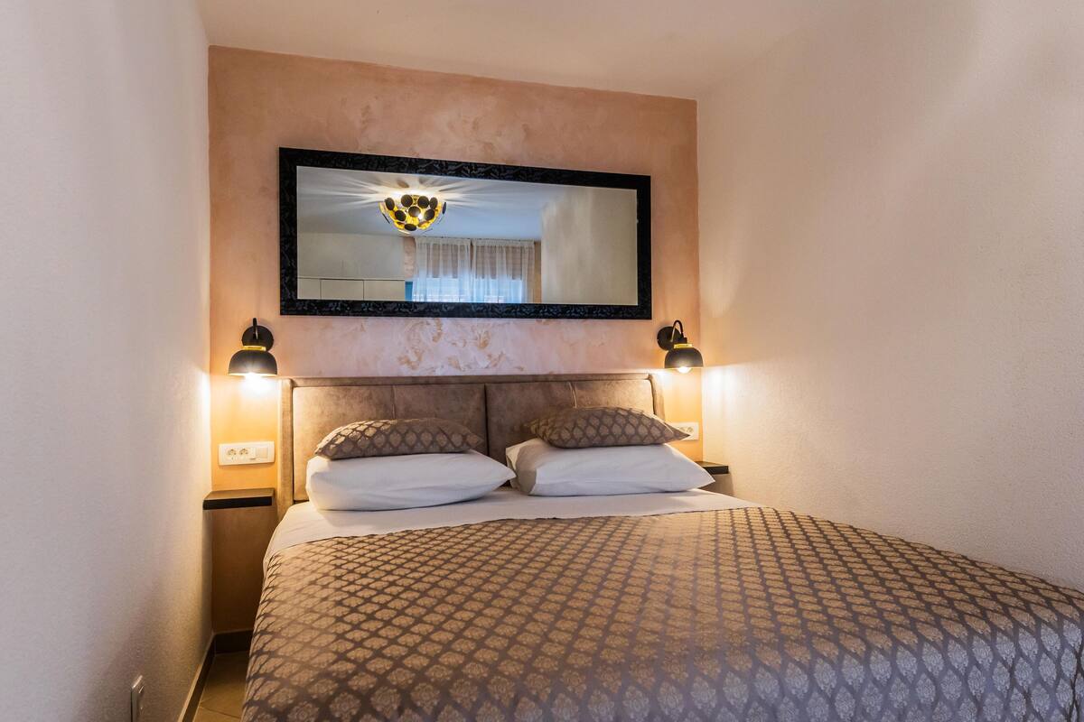 A hotel room with a bed, headboard, two bedside lamps, a large mirror, and a wall-mounted light fixture.