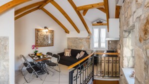 A cozy loft-style room with exposed wooden beams, a stone wall, a black sofa, a dining table with chairs, and a small kitchen area.