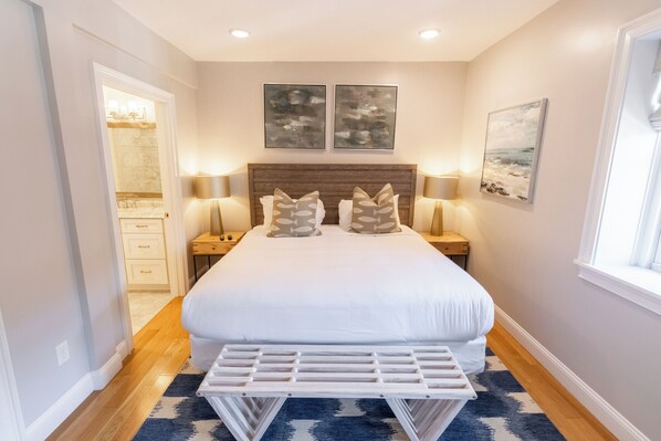 A bedroom with a large bed, two wall-mounted artworks, bedside lamps, and a view of the bathroom through an open door.