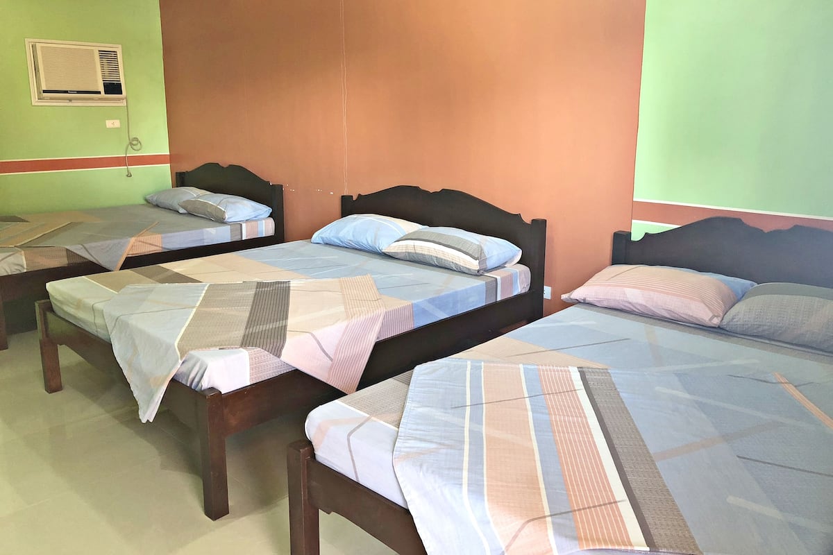 A room with four single beds, each with a wooden headboard and a striped bedspread. There is a wall-mounted air conditioning unit on the left wall.