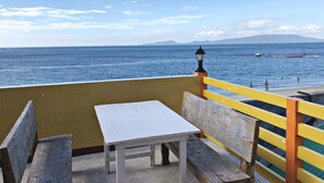 Une terrasse avec une table et des bancs donnant sur la mer, où l’on peut voir des bateaux sur l’eau et des montagnes au loin.
