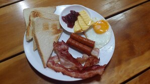 Une assiette avec des toasts, du bacon, des saucisses, un œuf au plat, de l’ananas et de la confiture.