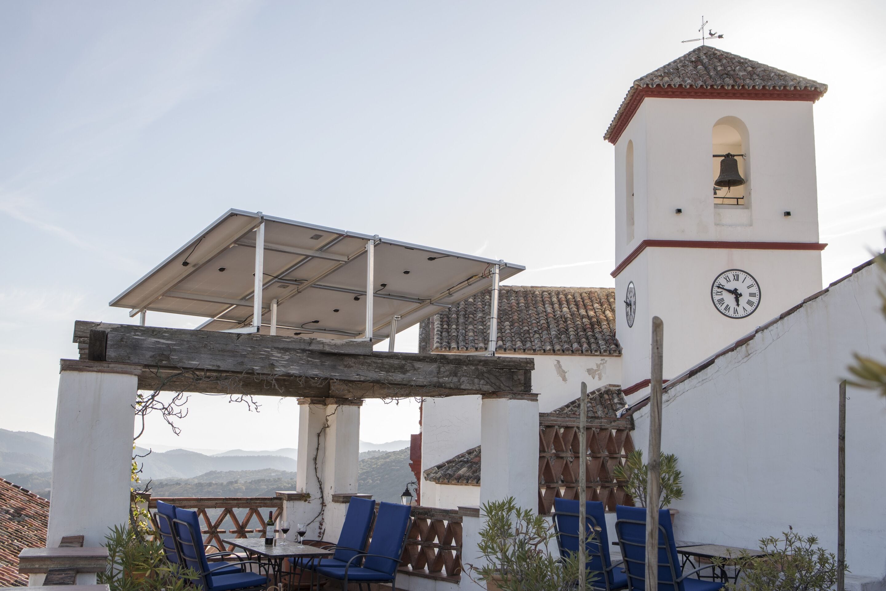 A terrace with blue chairs and a table, a white building with a bell tower, and a clock.
