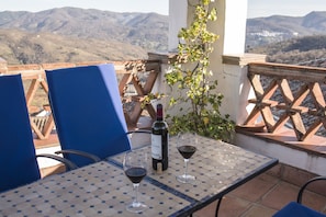 A table with a bottle of wine and two glasses, blue chairs, and a view of mountains. - Hotel Los Castaños (Cartajima)