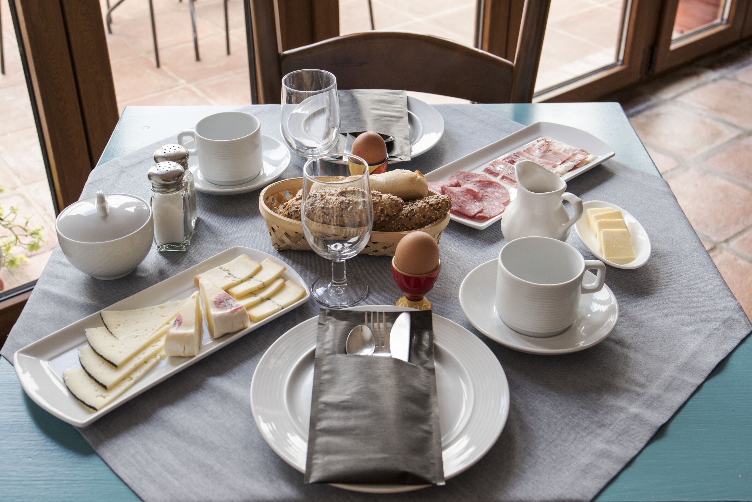 Une table de petit-déjeuner avec des assiettes remplies de nourriture, un panier de pain, une carafe de lait et un salier.