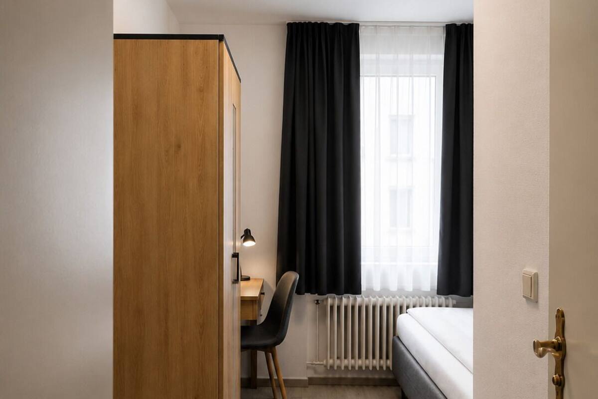 Kompakti hotellihuone, jossa on puinen vaatekaappi, työpöytä tuolilla, sänky ja ikkuna, jossa on verhot.