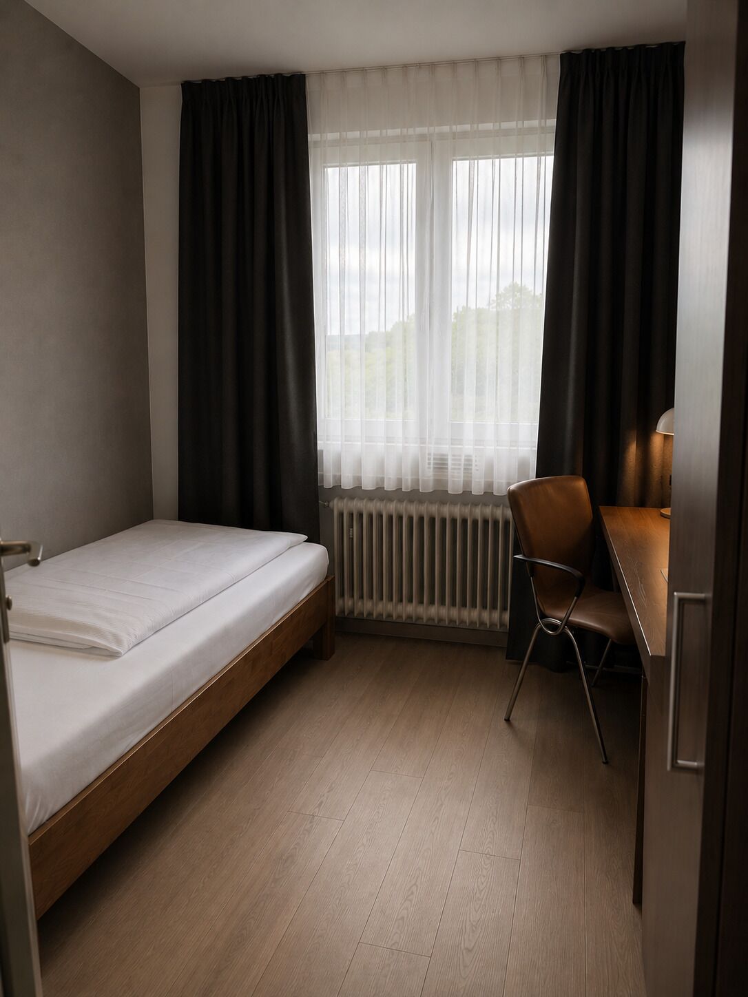 A single bed with a wooden frame, a desk with a chair, a radiator, and sheer curtains on a window.