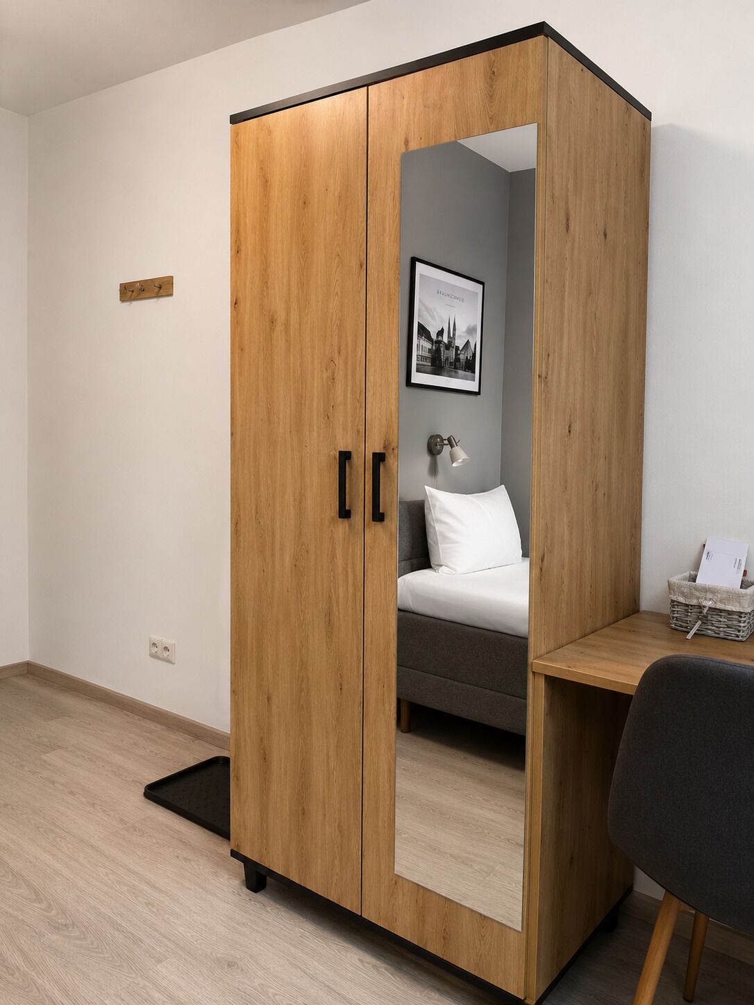 A room with a wooden wardrobe, a bed with white bedding, a desk with a chair, and a framed picture on the wall.