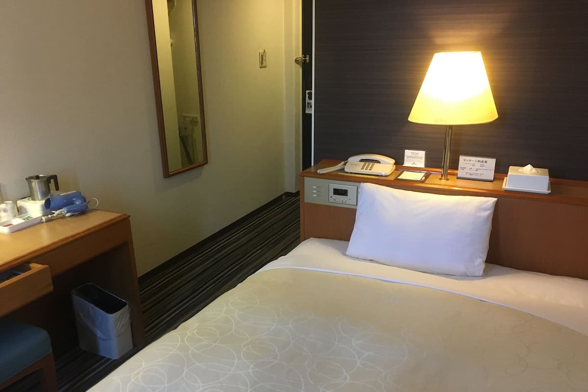 A hotel room with a bed, bedside table, lamp, phone, and a trash bin.