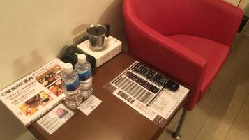 A hotel room with a brown table, a red chair, two water bottles, a kettle, a remote control, and a menu.