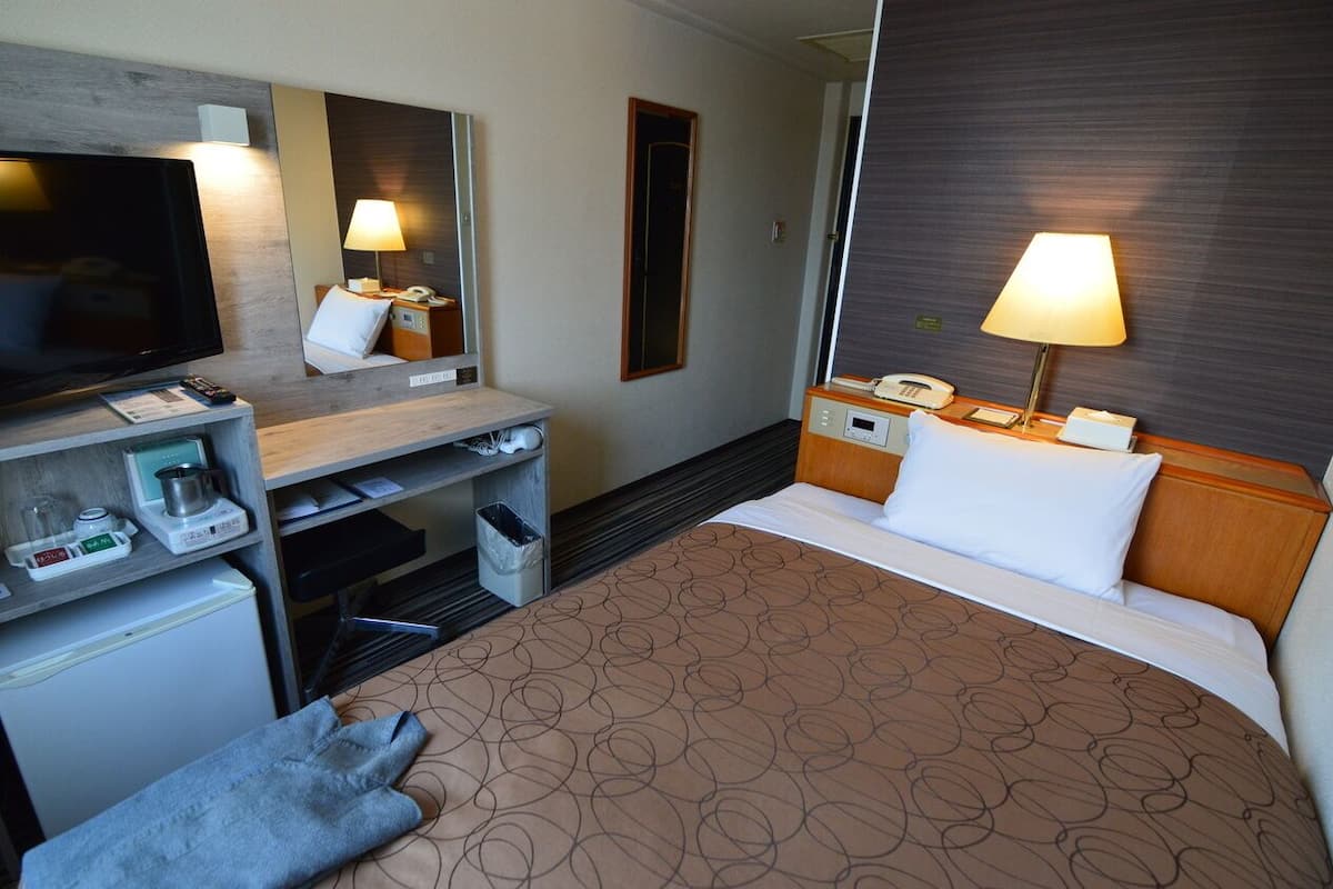 A hotel room with a bed, TV, desk, lamp, and a small refrigerator.