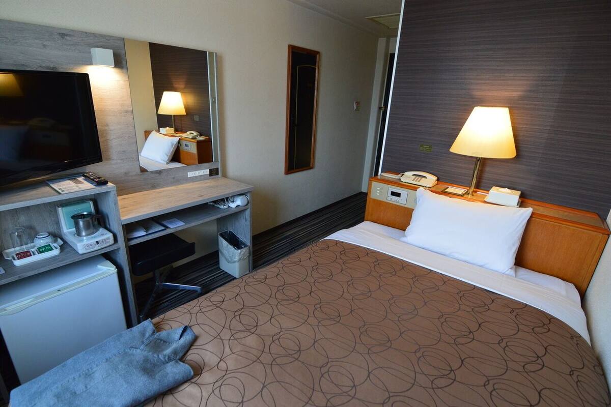 Habitación de hotel con cama, televisión, escritorio, lámpara y un refrigerador pequeño.