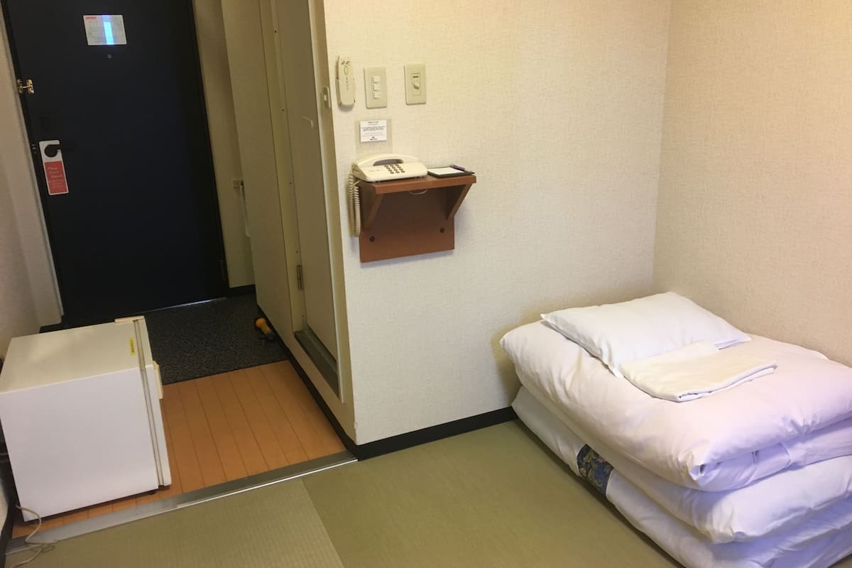 A small hotel room with a bed, a bedside table with a phone, a door, and a small white cabinet.