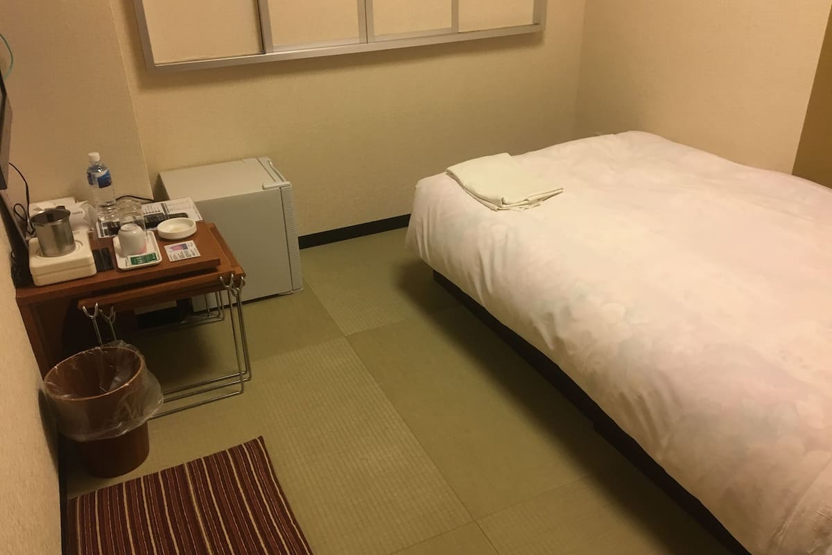 A hotel room with a bed, a small desk with a scale, a kettle, a cup, and a trash bin.