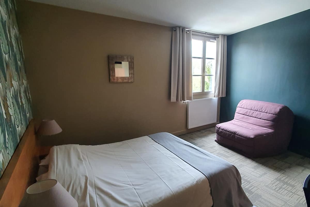 Een hotelkamer met een bed, een stoel, een raam met gordijnen, een radiator en een spiegel aan de muur.
