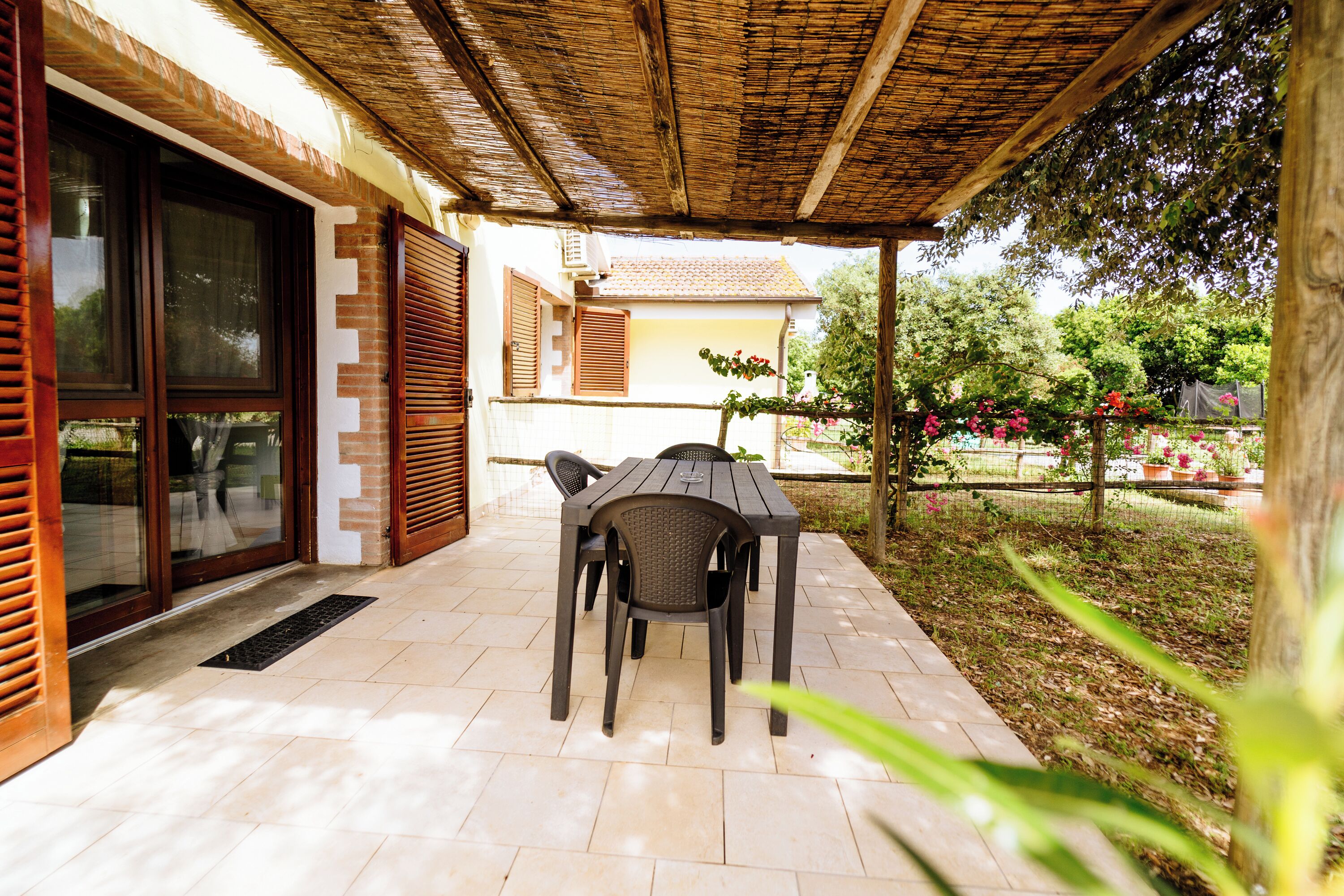 An outdoor patio with a table and chairs, a covered area with a wooden ceiling, and a view of a garden with trees and flowers.