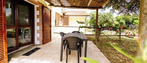 An outdoor patio with a table and chairs, a covered area with a wooden ceiling, and a view of a garden with trees and flowers.