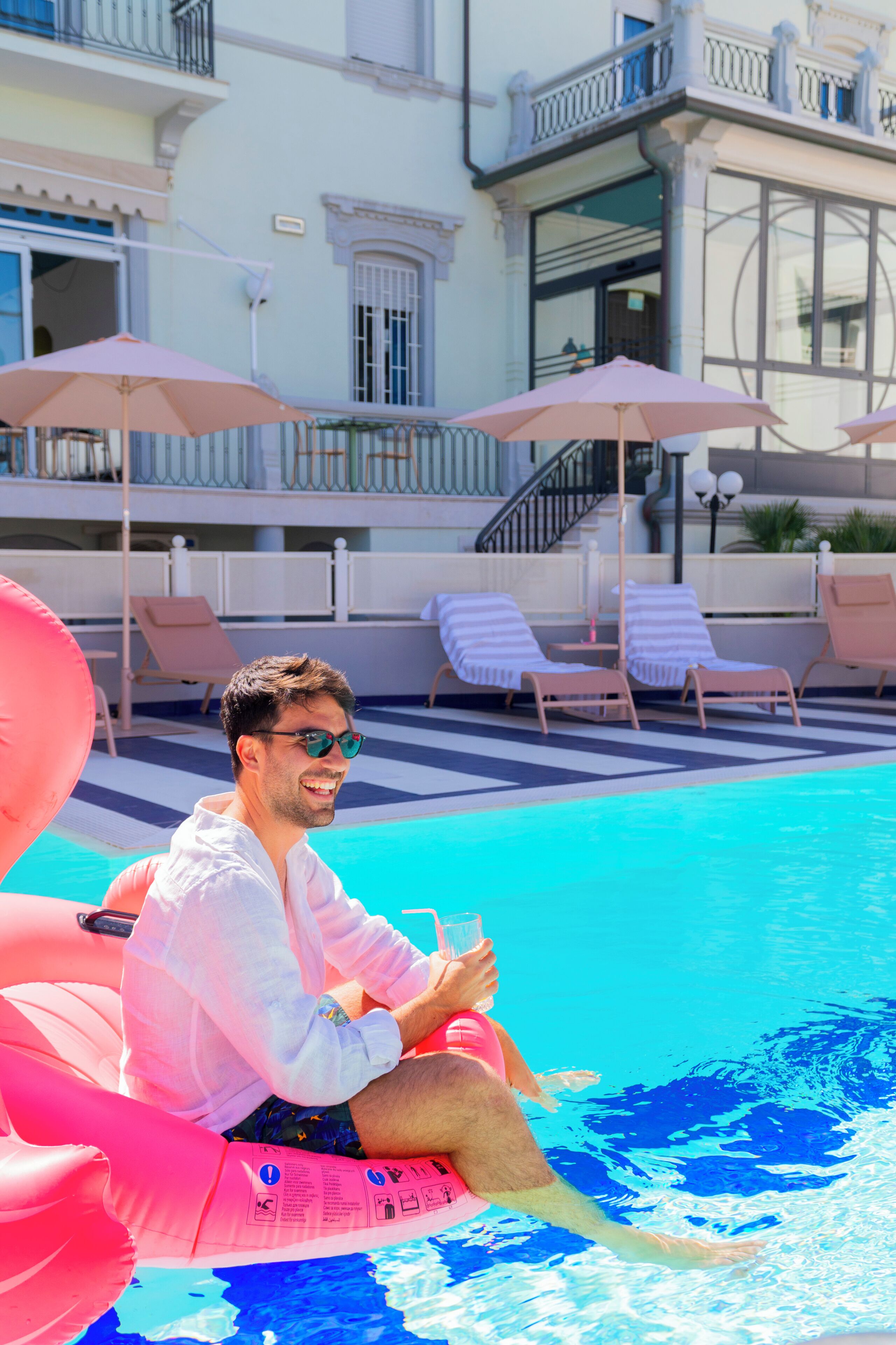 A pool area with lounge chairs and umbrellas, a person sitting on an inflatable float, and a building with balconies in the background.