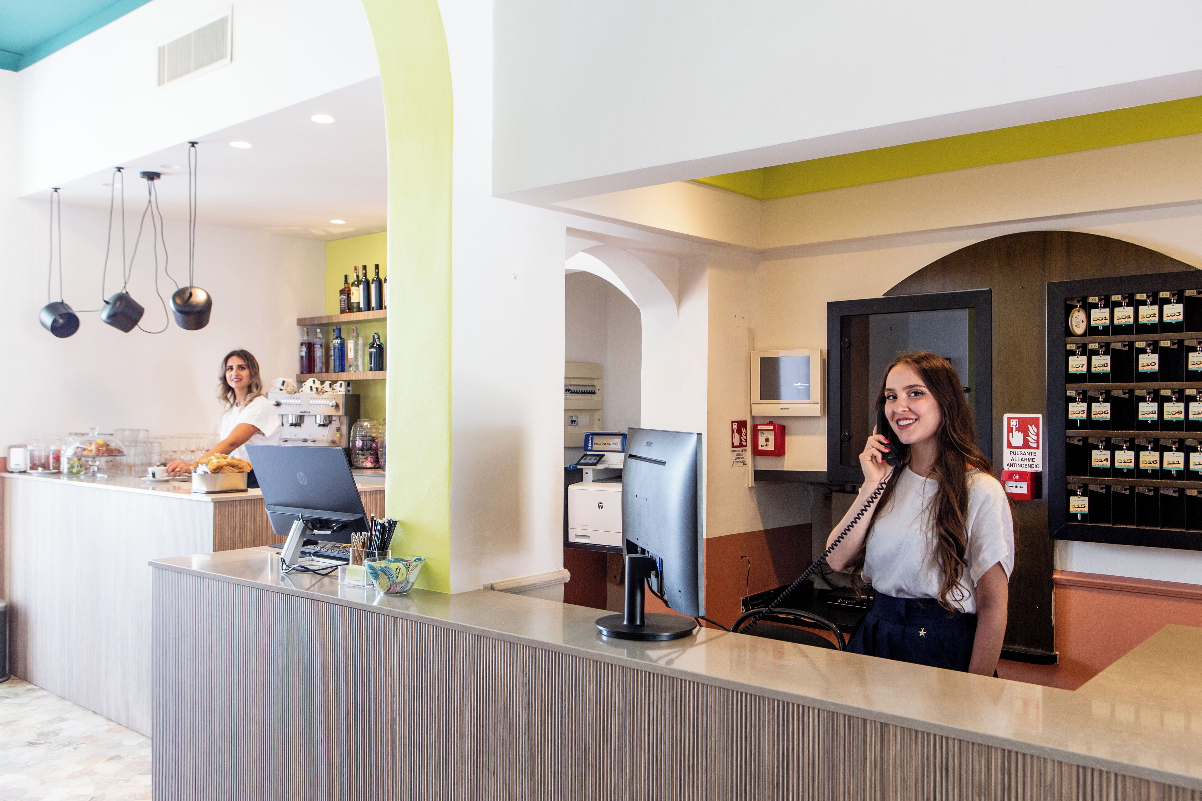 A hotel reception area with a counter, a computer, and a phone. There are shelves with bottles and a fire safety sign.