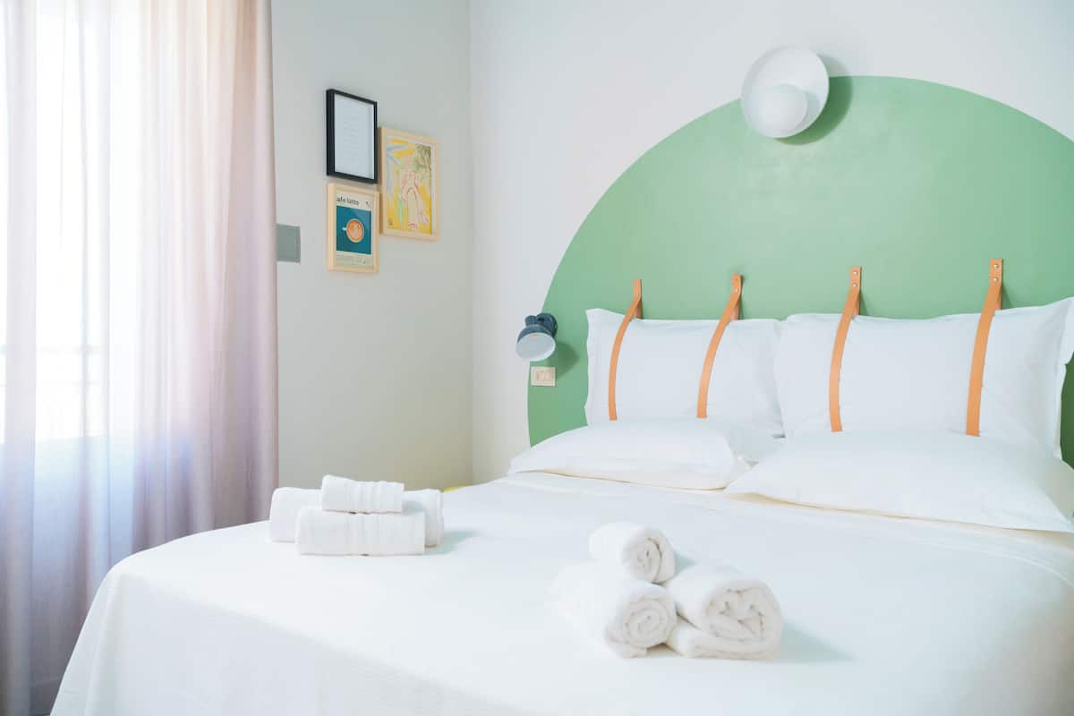 A neatly made bed with white linens and rolled towels, a green headboard, and a wall-mounted lamp.