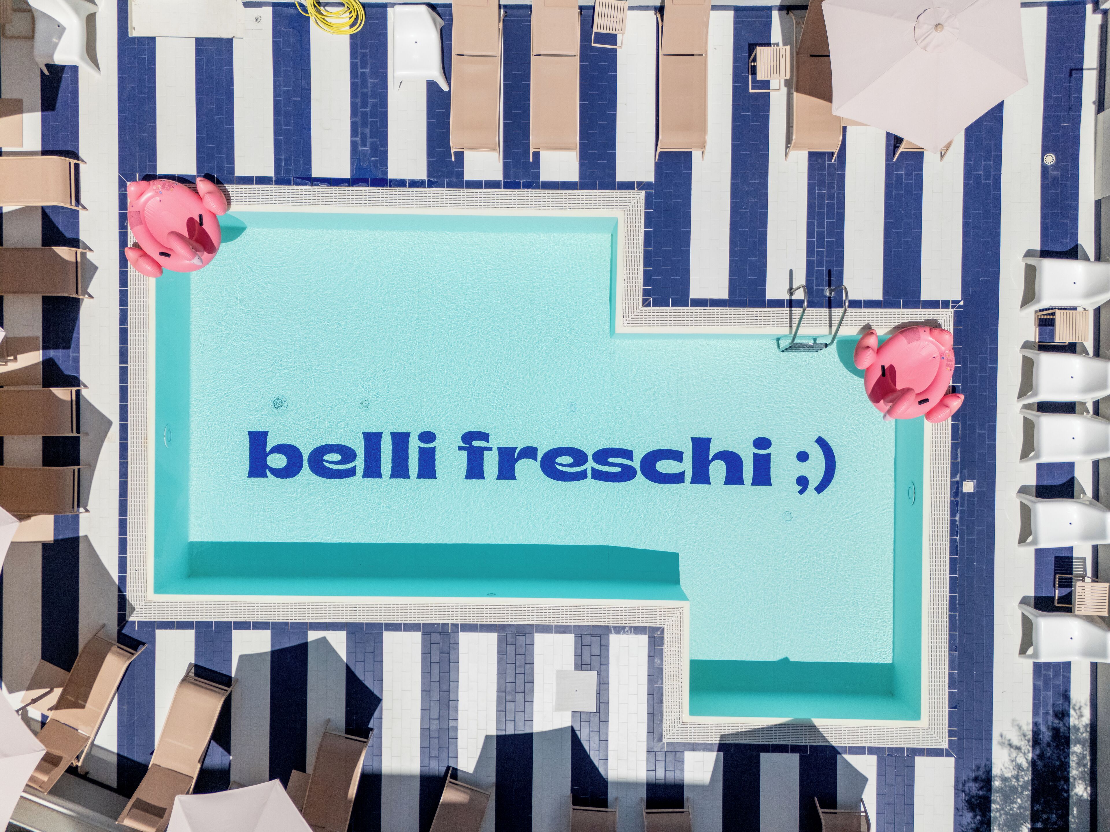 A pool area with a sign reading "belli freschi ;)" and poolside chairs and umbrellas.
