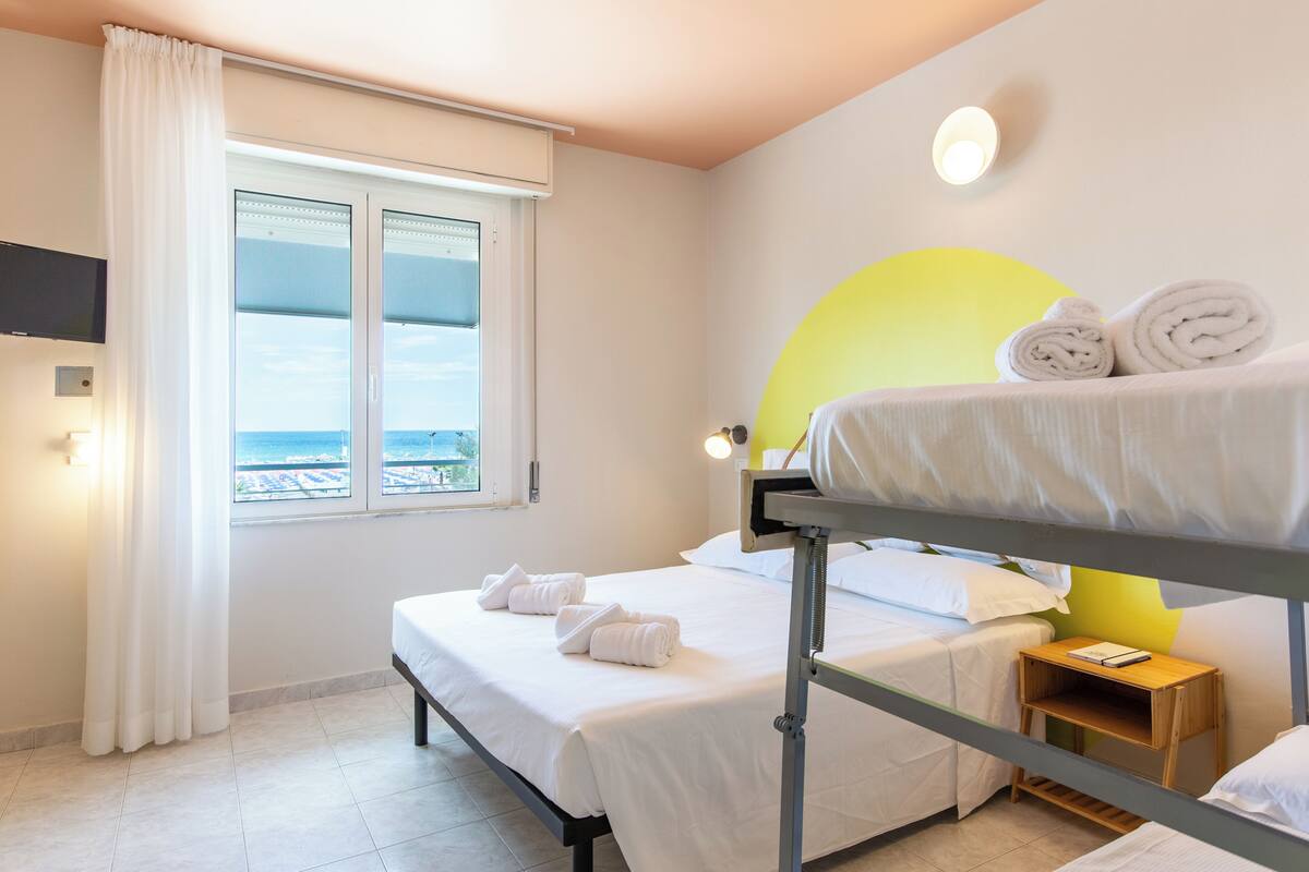 A hotel room with a bunk bed, a bedside table with a book, a window with curtains, and a wall-mounted TV.