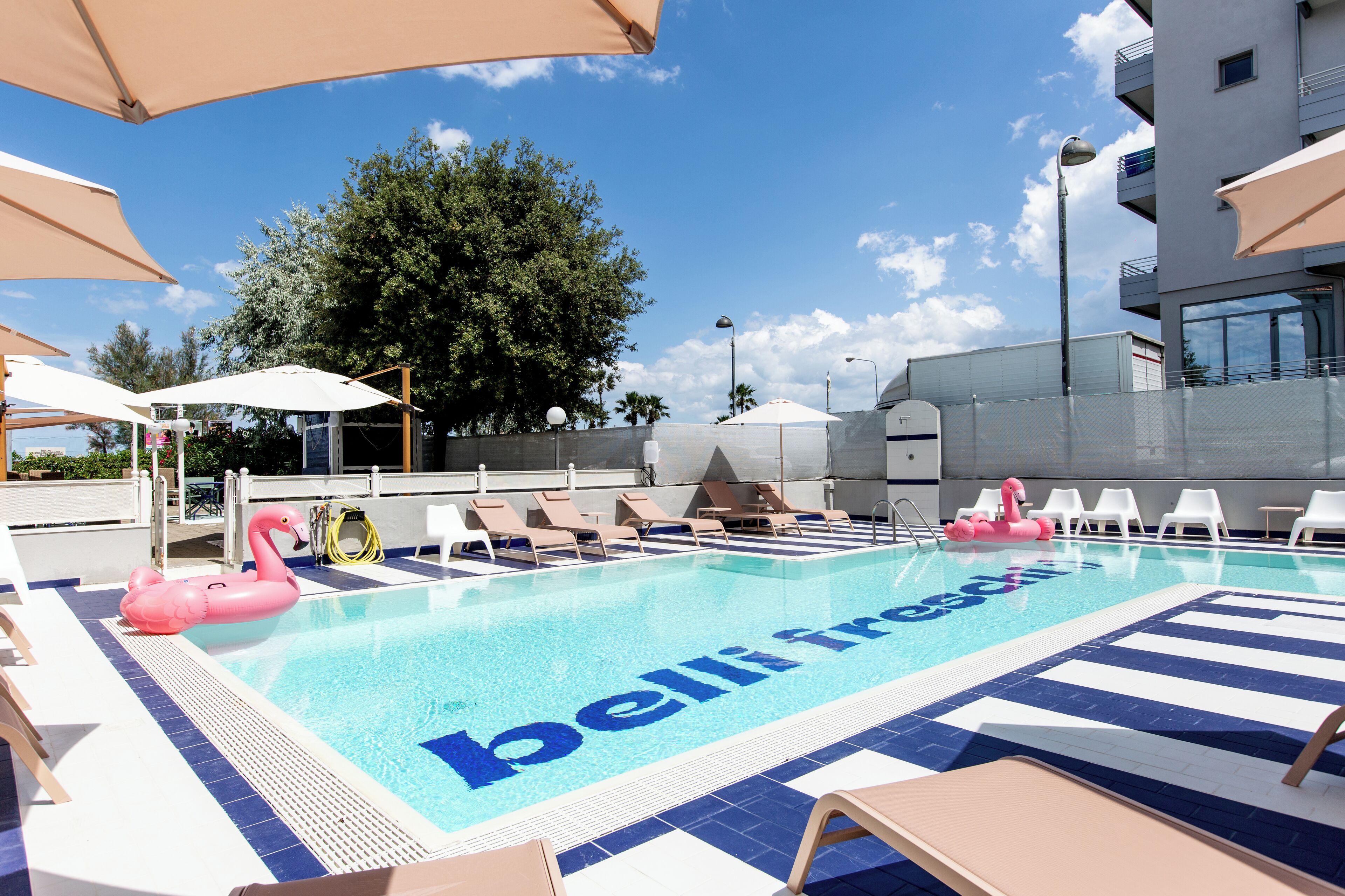 A swimming pool with lounge chairs and umbrellas, surrounded by a building with a balcony.
