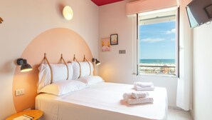 A bedroom with a bed, bedside lamps, a small table, and a window with a beach view. - Hotel Belvedere Rimini (Rimini)