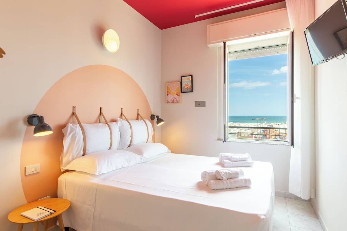 A bedroom with a bed, bedside lamps, a small table, and a window with a beach view.
