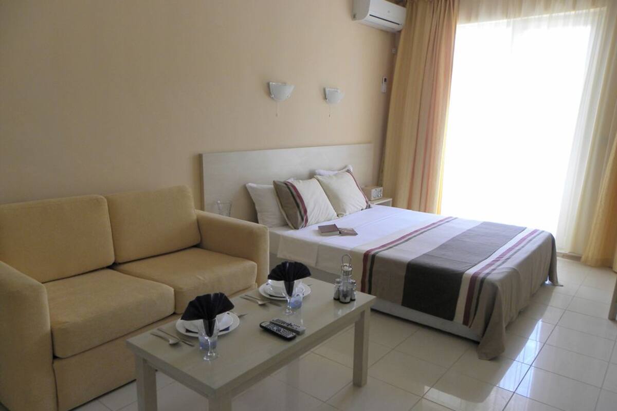 A hotel room with a bed, a sofa, a small table with a remote control, a glass, and a water bottle.