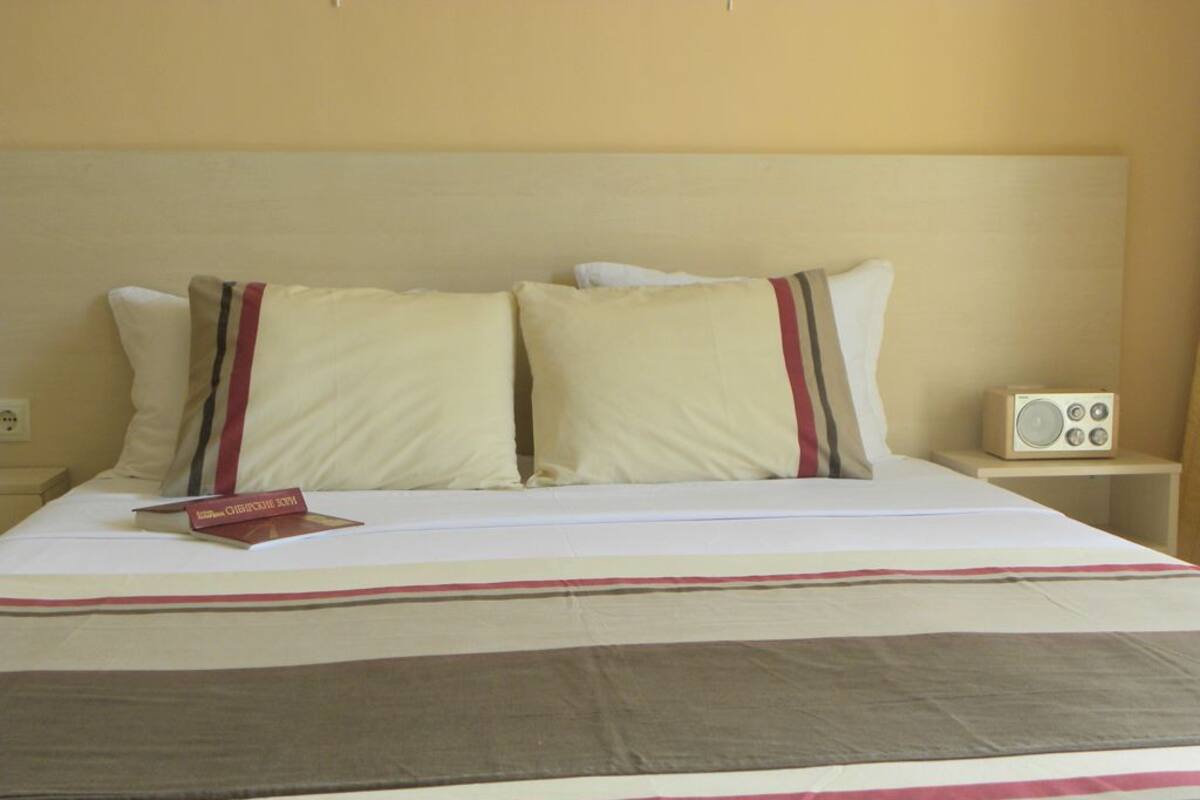 A hotel room with a bed, pillows, a bedside table with a radio, and a book on the bed.