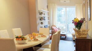 Un coin repas avec une table ronde, des chaises blanches, un panier de fruits et un vase de fleurs. On y trouve également un buffet en bois et une fenêtre avec des rideaux.