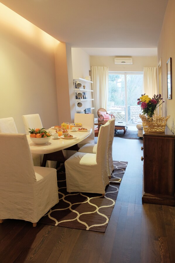 A dining area with a round table, white chairs, a fruit bowl, and a vase of flowers. There is a wooden cabinet and a window with curtains.